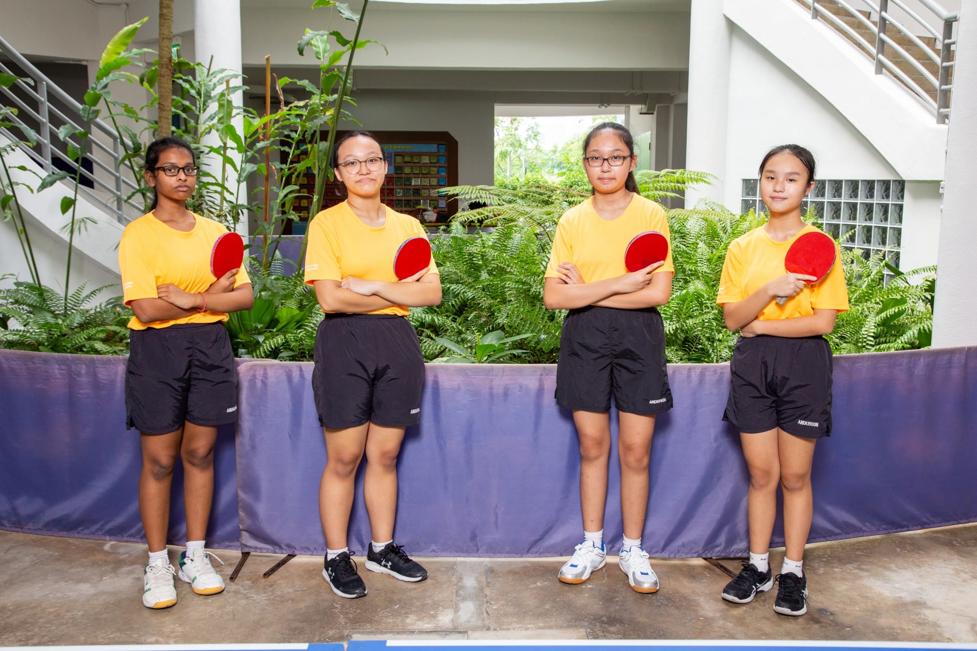 Four girls in yellow shirts and black shorts hold red table tennis paddles, posing together indoors near plants.